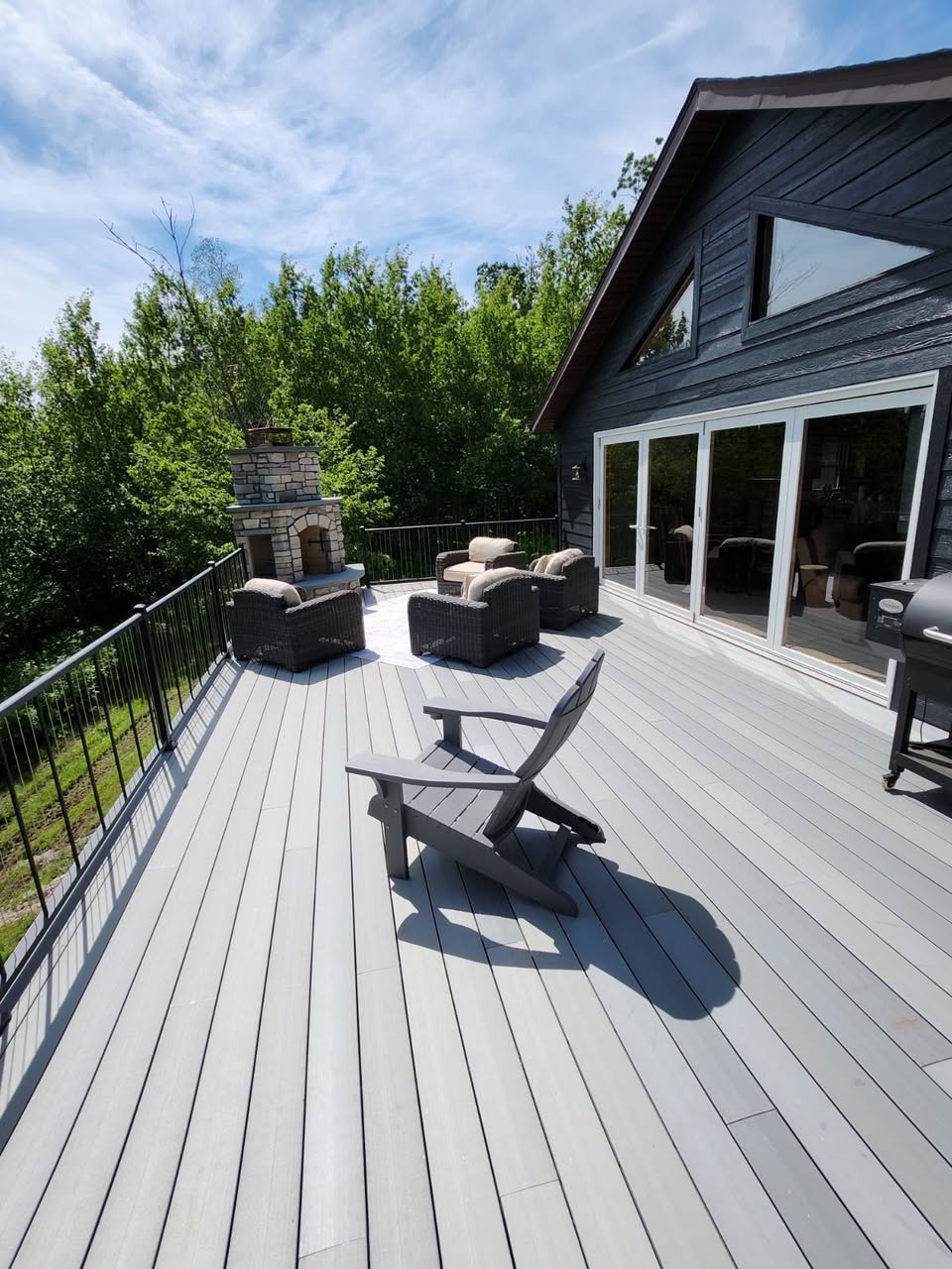 Large gray composite deck with black aluminum railings and a stone outdoor fireplace beside a dark-stained home by T & A Contracting